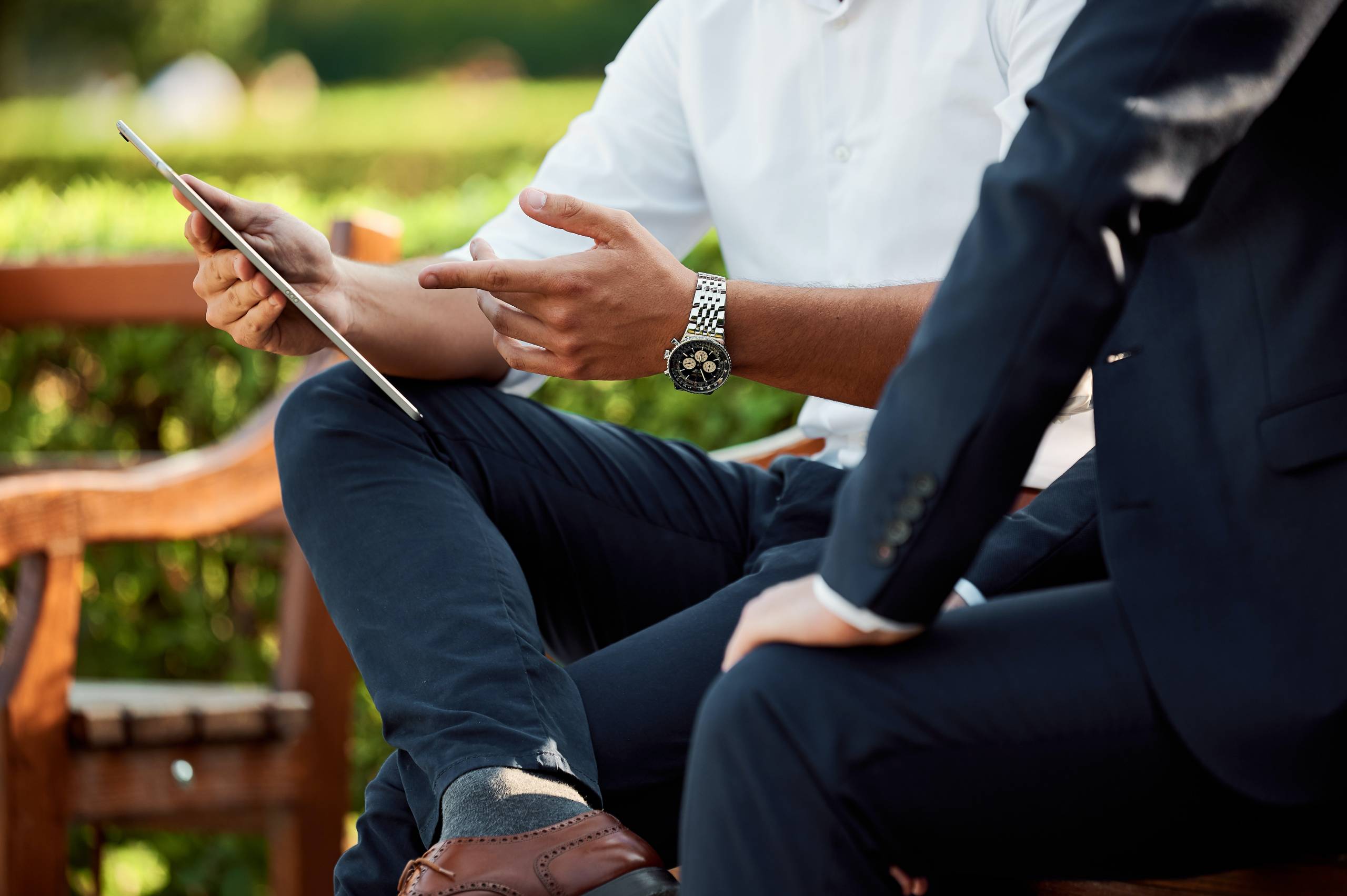 two-businesspeople-sit-on-park-bench-one-shows-ipad-screen-with-explanatory-gesture